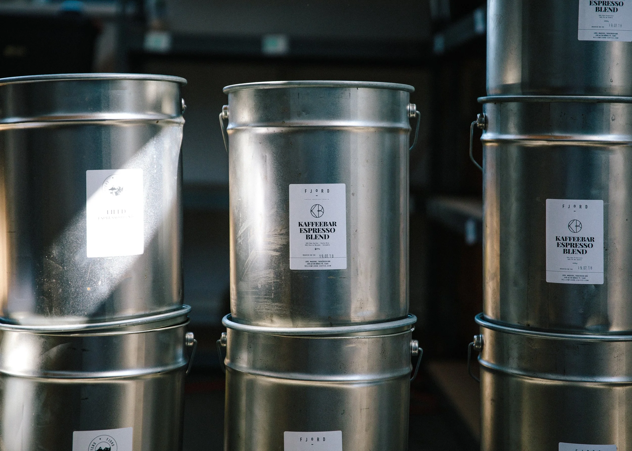 Stacked metal coffee cans labeled “Kaffebar Espresso Blend” in a roastery storage area.