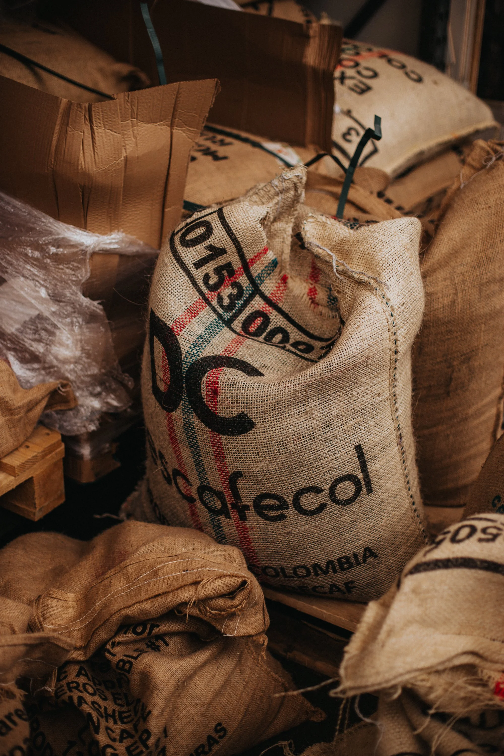 Burlap sacks marked “Coffee Colombia” stored in a warehouse with boxes and wrapping.
