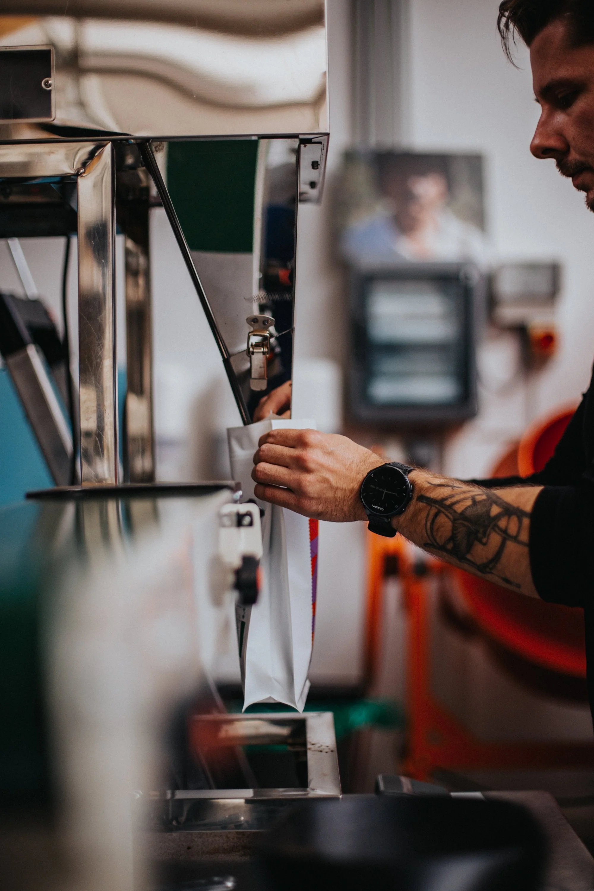 Tattooed roaster filling a paper bag with freshly roasted coffee beans in a Berlin roastery.