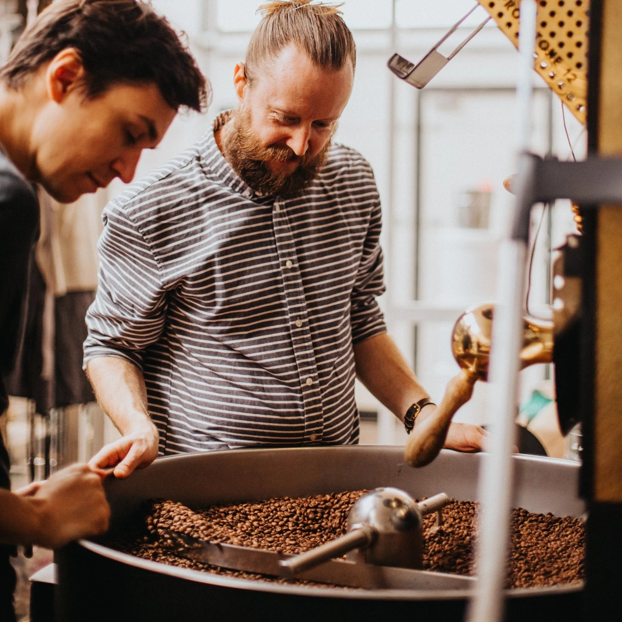 Two roasters inspecting coffee beans in a roasting machine, one with a scoop.