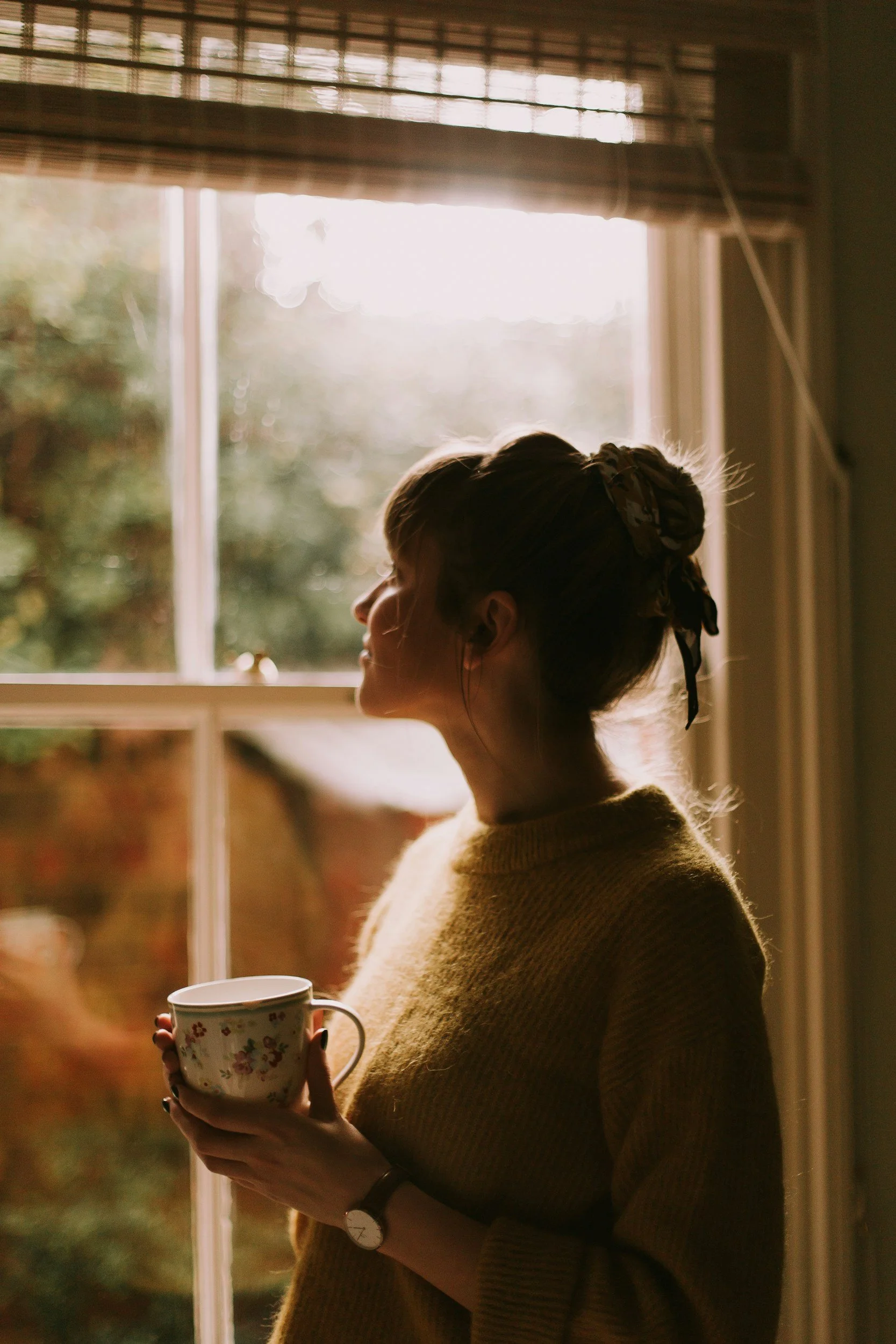 A woman standing by a window, holding a floral mug, while looking outside in warm sunlight.