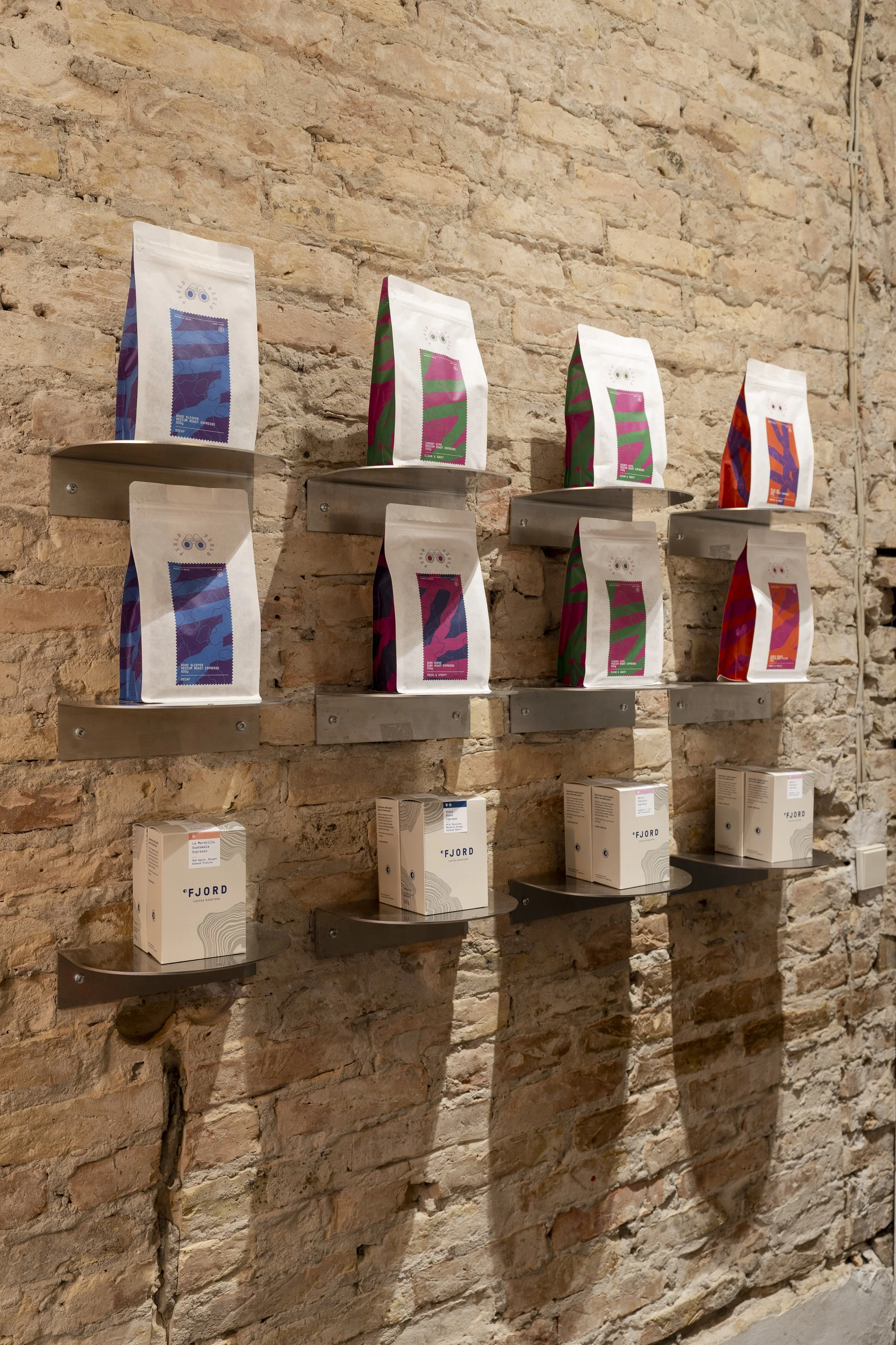 Display of colorful packaged products on metal shelves against a brick wall. The top row shows bags with vibrant abstract designs, and the bottom row contains white boxes with the label 'FJORD'.