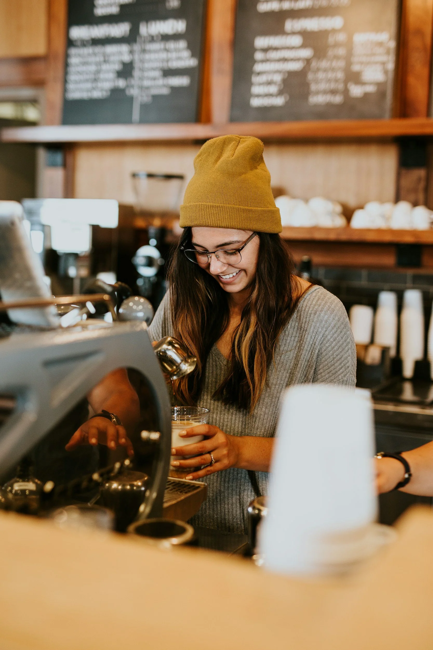 A woman with long brown hair, glasses, and a yellow beanie, smiling while making coffee at a coffee shop counter.